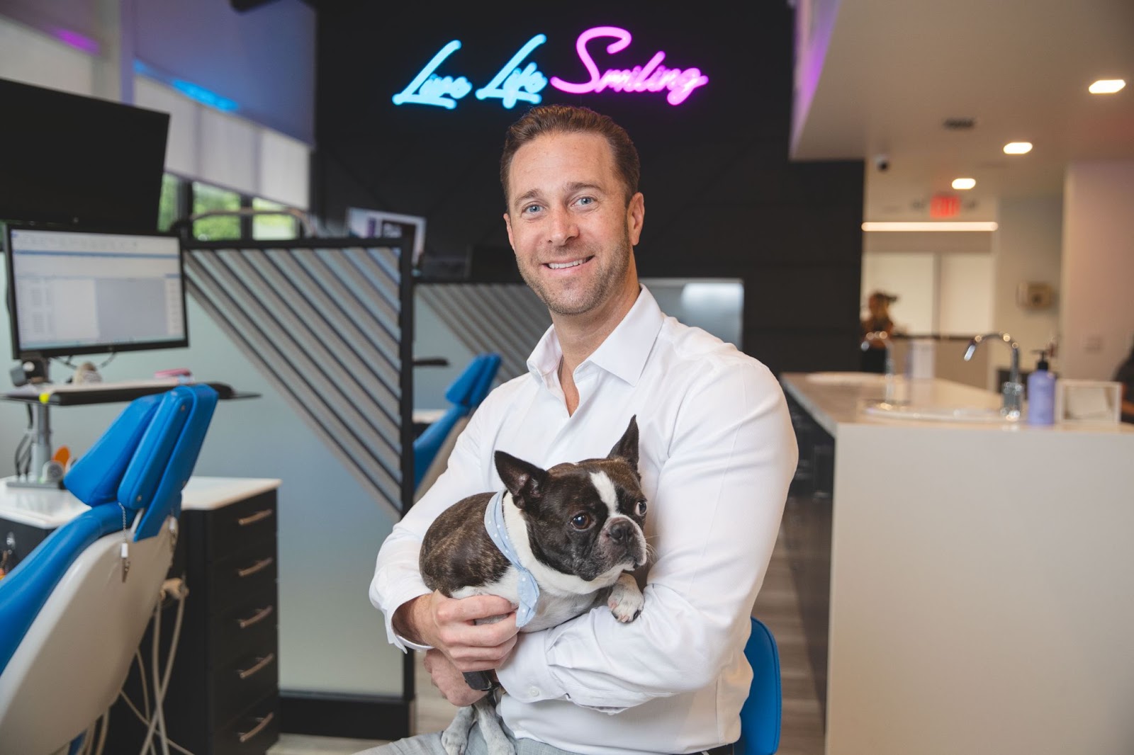 Orthodontist holding a French Bulldog in a modern orthodontic office with "Live Life Smiling" neon sign in the background.