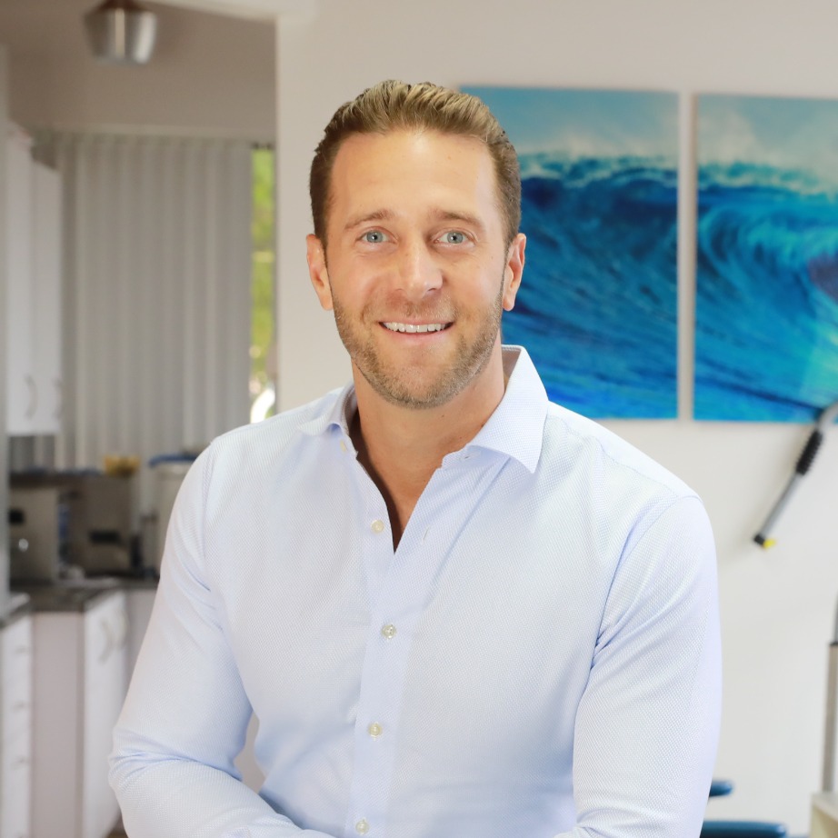 Dr. Ross, orthodontist at Kendall Orthodontics, smiling in a professional setting with dental equipment and ocean-themed artwork in the background.